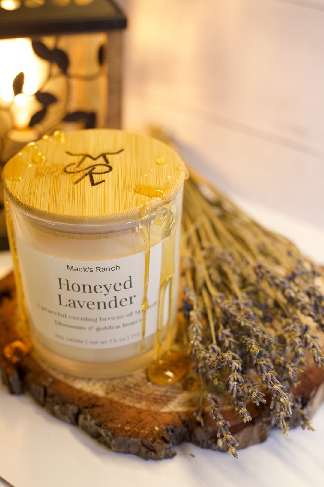 Honeyed Lavender candle with wooden lid and label with dripping honey on a wooden coaster with lavender flowers.